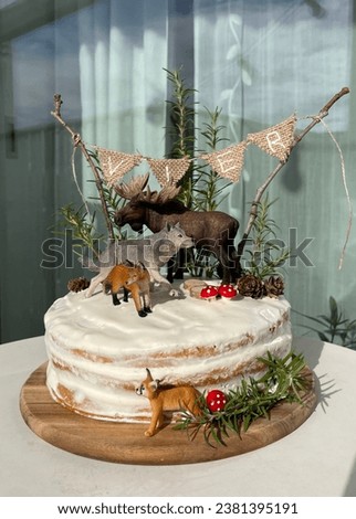 White Birthday cake on wooden plate with decorations of wild moose, wolf, fox and lynx standing in the forest among mushrooms and pine cones under the twine banner with letters VIER