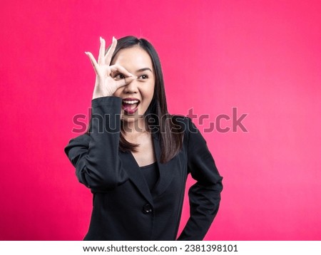 Portrait of an Asian Indonesian woman wearing a black work shirt, posing in agreement with an OK sign in front of her eyes. Isolated against a magenta background.