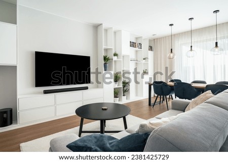 Modern living room with dining area in clean design with builtin furniture and window.There're large TV with sound bar,coffee table,many decorative plants,massive table with chairs and ceiling lights. Royalty-Free Stock Photo #2381425729