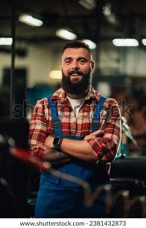 A portrait of a young factory worker with a tattoo on the forearm and a beard, wearing blue overalls and red and white plaid shirt. He is smiling with arms crossed. Factory setting.