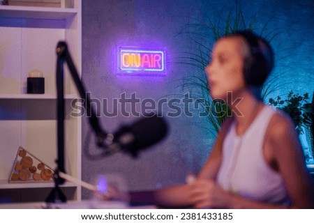 Selective focus on the neon "on air" sign on the wall in a small recording and podcasting studio with a burred girl wearing headphones in the foreground. A radio sign on the wall with a blurry girl.