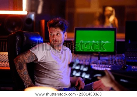 A picture of a music producer sitting in front of a mixing console and a monitor in a music studio. He has a tattooed arm and fashionable hair. In the background, there is a singer in the silent room.