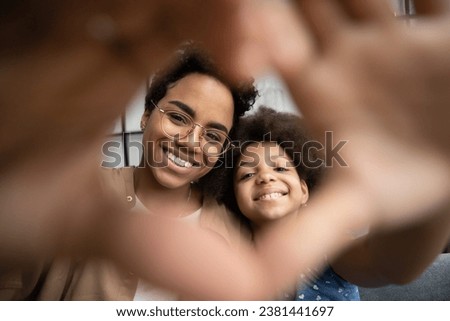 Happy young African American mommy and cute pre teen daughter kid looking at camera through finger heart frame, joining hands, showing symbol of love, tenderness, gratitude, affection. Close up Royalty-Free Stock Photo #2381441697