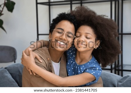 Peaceful happy Black mom and cute fuzzy haired daughter girl hugging with love and tenderness, smiling with closed eyes, enjoying warm moment at home. Motherhood, family leisure