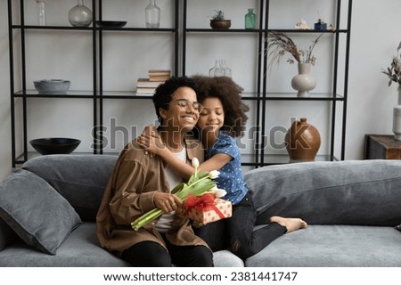 Happy loving African pre teen kid embracing grateful young mom holding present box, tulip flowers, relaxing on couch, smiling. Mum and kid celebrating mothers day, birthday, festive event Royalty-Free Stock Photo #2381441747