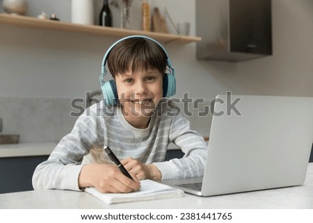 Happy positive early teen gen Z schoolchild wearing big stylish headphones, studying at home, using laptop, writing notes, looking at camera, smiling. School boy with dental braces head hot portrait