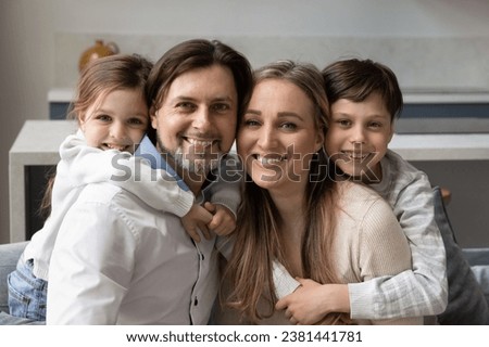 Happy cheerful couple of parents piggybacking sweet little daughter girl and teen son kid, hugging, sitting close on home sofa, looking at camera with toothy smiles. Family head shot portrait