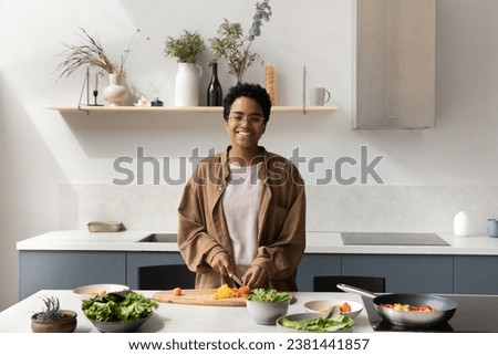Happy African chef girl cooking vegetarian meals in home kitchen, slicing fresh vegetables at table, preparing organic natural ingredients, making dinner, looking at camera, smiling