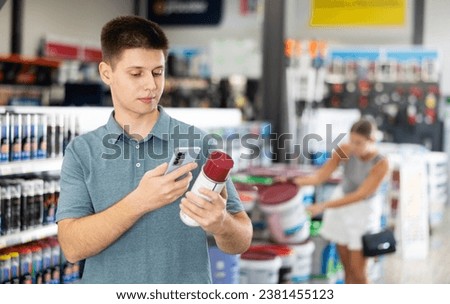 Guy chooses picks up buys aerosol paint varnish solvent in construction store. Young man visitor carefully studies information on product label and photographs packaging of product with phone