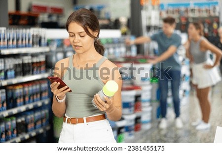 Girl young woman chooses picks up buys aerosol paint varnish solvent in construction store. Woman visitor carefully studies information on product label and photographs packaging of product with phone