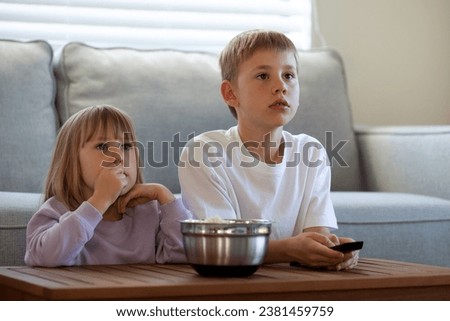 Cute brother and sister are sitting at the table with a bowl of popcorn and enthusiastically watching TV. The concept of children's leisure, pastime.