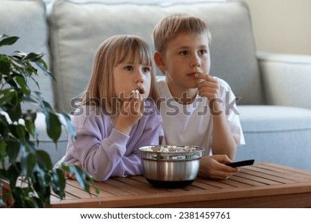 Cute brother and sister are sitting at the table with a bowl of popcorn and enthusiastically watching TV. The concept of children's leisure, pastime.