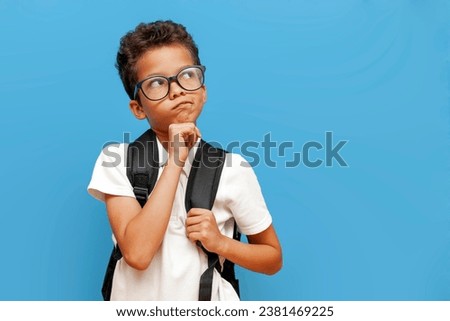 pensive african american teenager boy in glasses with backpack thinks and plans on blue isolated background, curly schoolboy imagines daydreaming and looks away Royalty-Free Stock Photo #2381469225