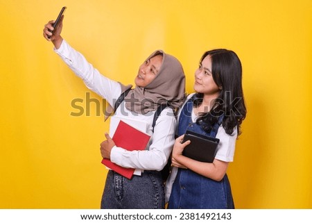 Cheerful student girls holding book and do selfie on mobile phone over yellow background