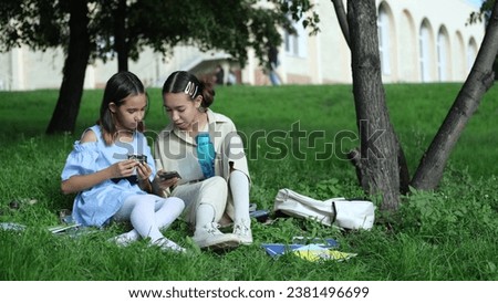 Happy sisters watch a video, take a selfie or talk on a video call using a smartphone outside in the park on a summer day.