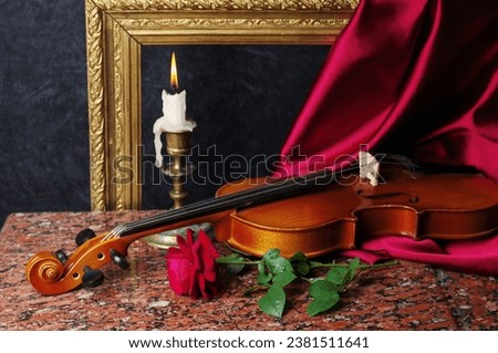 A burning candle, a violin with a bow and a red rose against the background of a picture frame and burgundy drapery.