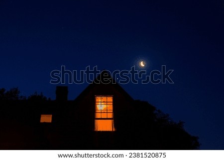 Mysterious gabled house silhouette with lit window, crescent moon and night sky. 