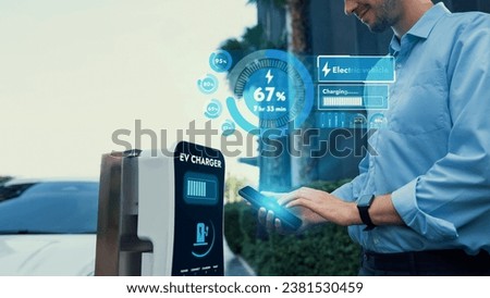 Businessman checking battery status hologram from EV smartphone application while his EV car recharging from charging station in residential area. Futuristic clean sustainable EV tech. Peruse