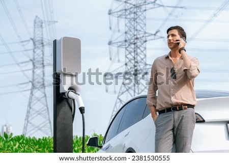 Man talking on the phone while recharge EV car battery at charging station connected to power grid tower electrical as electrical industry for eco friendly car utilization.Expedient Royalty-Free Stock Photo #2381530555