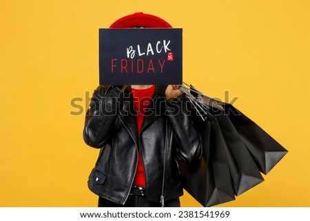 Young woman wearing casual clothes red hat hold shopping paper package bags cover face card sign with Black Friday written text inscription isolated on plain yellow background. Sale buy day concept