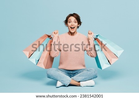 Full body young woman she wear beige knitted sweater casual clothes sitting hold in hand paper package bags after shopping isolated on plain blue background studio. Black Friday sale buy day concept