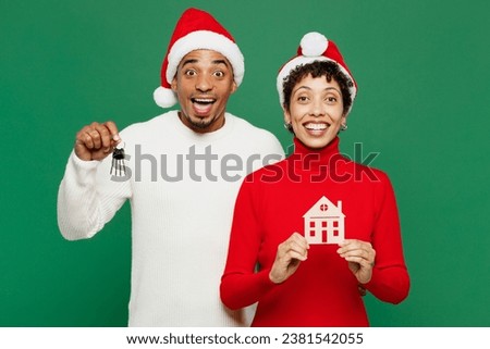 Merry cool young couple friends man woman wear red casual clothes Santa hat posing hold keys wooden mock up of house isolated on plain green background. Happy New Year 2024 Christmas holiday concept