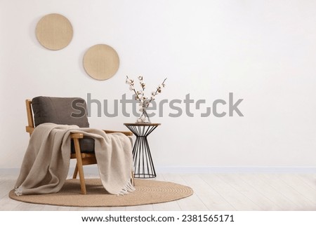 Comfortable armchair, blanket, side table and vase with cotton branches near white wall indoors. Space for text Royalty-Free Stock Photo #2381565171