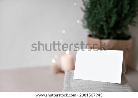 Blank paper card mockup, sweaters on table, blurred defocused candle and garland lights, juniper in pot on background. Empty neutral beige wall copy space. Christmas postcard or invitation template