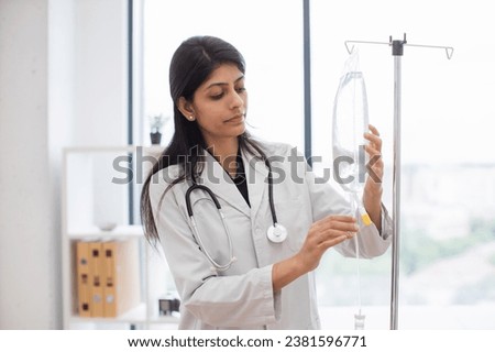 Adult female doctor in white lab coat standing near medical dropper and controlling liquid medication. Indian woman preparing infusion drip for intravenous treatment system. Royalty-Free Stock Photo #2381596771