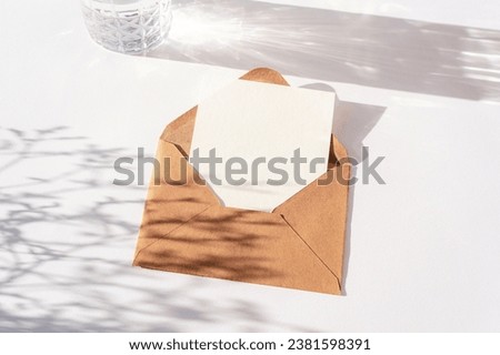 Envelope with blank card, glass of water and flowers shadow on white table. Top view, flat lay, mockup.