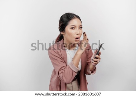 A young beautiful Asian woman employee wearing cardigan and holding her phone is shouting and screaming loud with a hand on her mouth, isolated by white background. Royalty-Free Stock Photo #2381601607