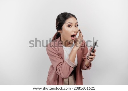 A young beautiful Asian woman employee wearing cardigan and holding her phone is shouting and screaming loud with a hand on her mouth, isolated by white background. Royalty-Free Stock Photo #2381601621