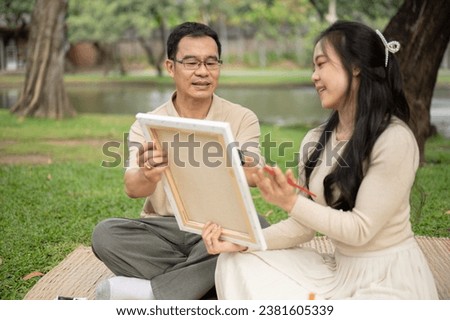 A lovely and cheerful Asian girl enjoys picnicking in a green park with her dad, spending time to create and paint a picture on a canvas together. Happy family and leisure concepts