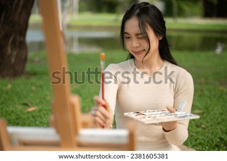A beautiful and creative young Asian female artist is measuring her picture with a paintbrush, enjoying painting on a canvas while relaxing in a green park.
