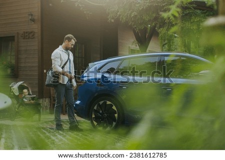 Portrait of a man charging electric car in front of his house, plugging the charger into the charging port. The man is unplugging charger from the fully charged car before going to the office. Royalty-Free Stock Photo #2381612785