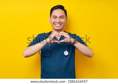 Smiling professional young Asian man doctor or nurse wearing a blue uniform and stethoscope showing heart shape with hands isolated on yellow background. Healthcare medicine concept