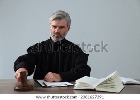Judge with gavel, papers and book sitting at wooden table against light grey background