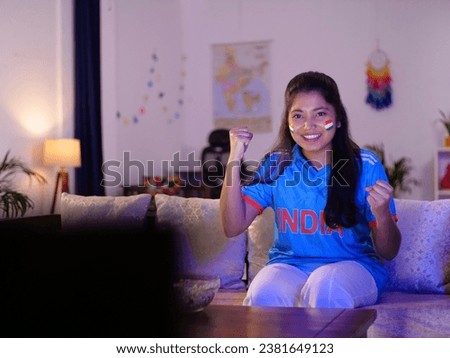 Young Indian girl celebrating Indian cricket team's victory - watching live cricket match on TV, famous sport. Female Indian cricket fan wearing an Indian jersey - excitement, cheering for team, fe...