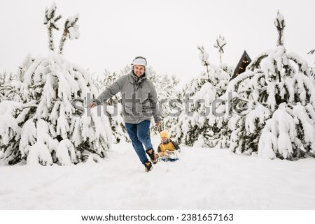 Father pulling little son on winter day. Children are rolling down hill on sledge in forest. Happy funny child with dad ride sled on snowy road in mountains. Family walks during snowfall in park.