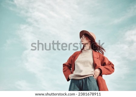 Confident teenage girl in red coat and hat standing against sky and looking away. Low angle view. Royalty-Free Stock Photo #2381678911