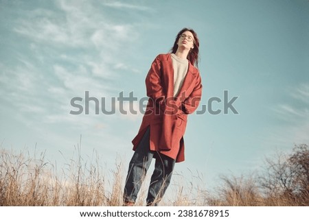 Teenage girl in a red coat stands against the sky with her hands in pockets. Royalty-Free Stock Photo #2381678915