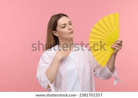 Beautiful woman waving yellow hand fan to cool herself on pink background