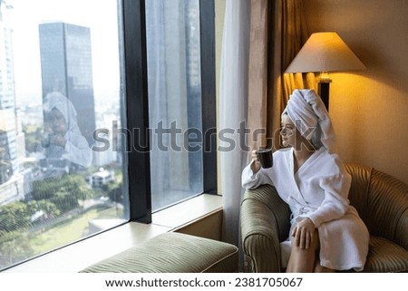 Young woman drinking morning coffee, wearing white bathrobe and towel on the head, looking at cityscape through the window in luxury penthouse apartment or hotel room Royalty-Free Stock Photo #2381705067