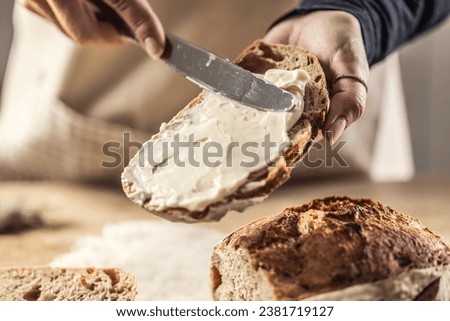 A woman makes delicious bread, spreads cream cheese with a cutlery knife - Close up. Woman hands spreading cream cheese on bread slice. Royalty-Free Stock Photo #2381719127