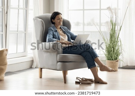 Middle-aged woman relax on comfortable armchair in living room with laptop, watch movie on digital streaming on-line services, spend time on internet, make order, enjoy leisure and modern tech usage Royalty-Free Stock Photo #2381719473