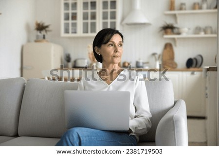 Serious focused mature female sit on sofa with notebook on laps, looks aside, thinking on text, search ideas for new post. Blogging, older generation and modern tech usage for study, work or purchases Royalty-Free Stock Photo #2381719503