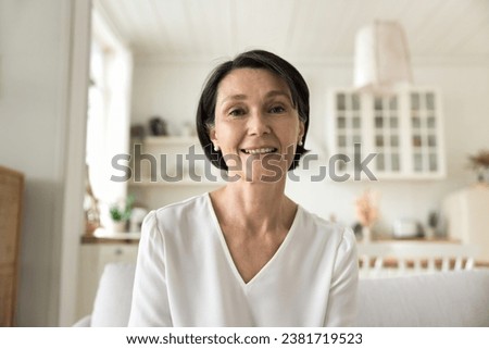 Attractive middle-aged woman smile looks at camera sit on sofa, having videoconference remote talk to family or grown-up children living abroad, enjoy communication using modern tech, profile picture