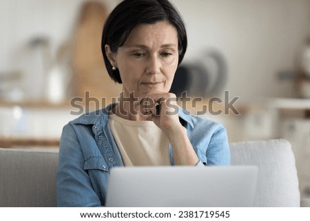 Concerned middle-aged woman looks at laptop screen, read e-mail, thinking on unpleasant news, learn new software, having lack of understanding of modern notebook usage sit on sofa at home, close up Royalty-Free Stock Photo #2381719545