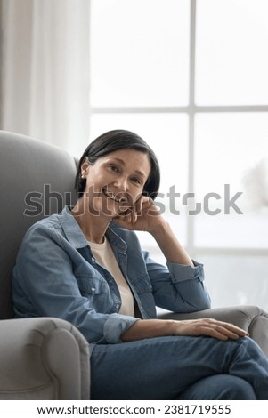 Good-looking middle-aged female in casual denim clothes posing staring at camera seated on modern, soft armchair alone at home, enjoy carefree rest, having attractive appearance and optimistic mood Royalty-Free Stock Photo #2381719555