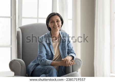 Middle-aged pretty woman in denim shirt and jeans posing for camera seated on modern armchair, smile enjoy carefree retired life, weekend leisure and relaxation at home, generation X female portrait Royalty-Free Stock Photo #2381719557